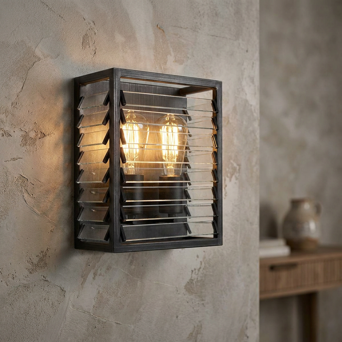 Industrial gunmetal wall light with glass louvre slats and two glowing Edison bulbs on a textured wall.