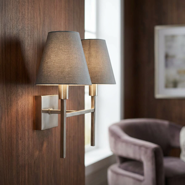 Brushed nickel two-light wall sconce with grey fabric shades mounted on a wood-paneled wall.