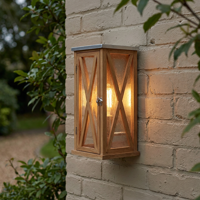 Natural oak outdoor wall lantern with X-frame detailing and a glowing bulb, mounted on a light brick wall.