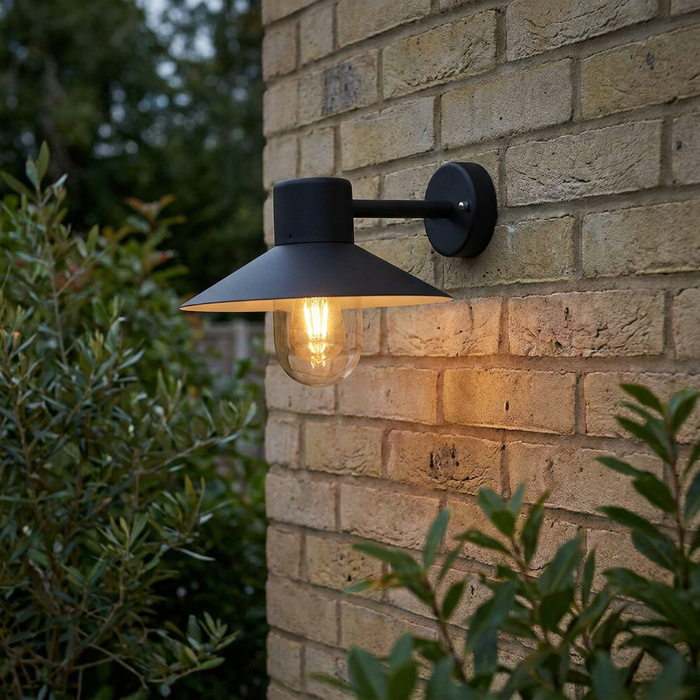 Black outdoor wall light with a conical shade and clear glass bulb mounted on a brick wall in a garden setting.