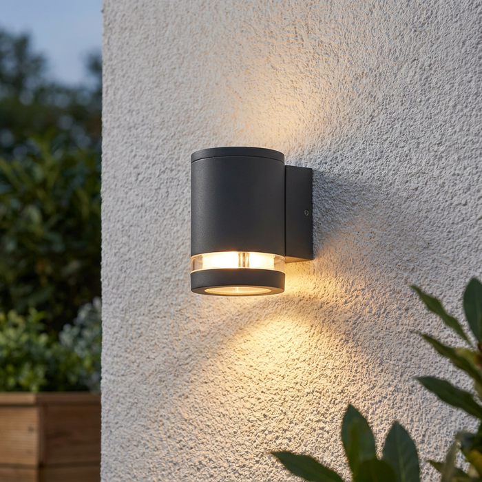 A cylindrical grey outdoor wall light illuminates a textured white wall with a warm downward glow.