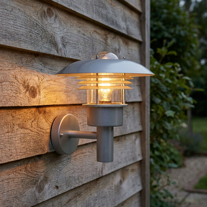 Modern silver outdoor wall light with tiered discs and a glowing bulb mounted on a rustic wooden fence.