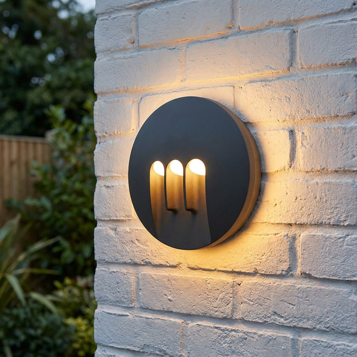 Circular graphite LED wall light with three vertical cutouts mounted on a white painted brick exterior wall.
