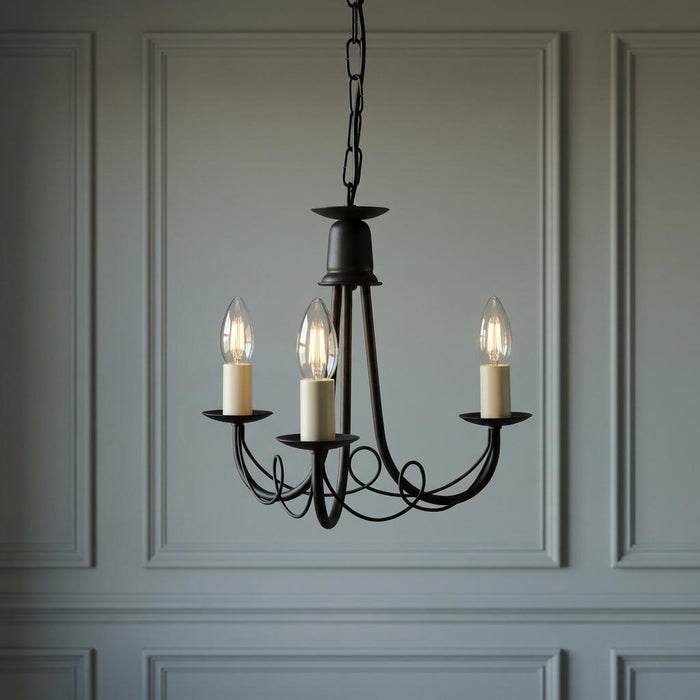 Black three-light scroll chandelier with lit candle bulbs against a light grey decorative paneled wall.