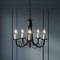Elstead Minster 5-light black chandelier with lit candle bulbs against a dark blue paneled wall.