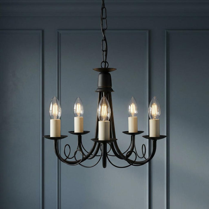 Elstead Minster 5-light black chandelier with lit candle bulbs against a dark blue paneled wall.