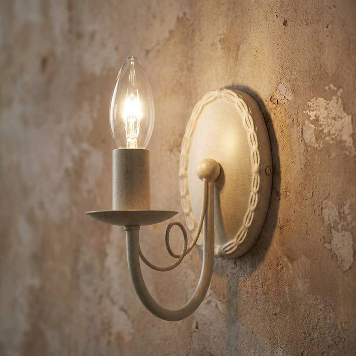 Ivory candle-style wall sconce with an ornate oval backplate, illuminated against a distressed wall.