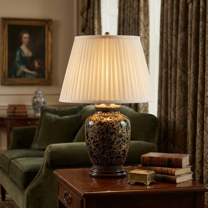 Lit ceramic lamp with black and gold floral patterns and a pleated shade sitting on a wooden side table.