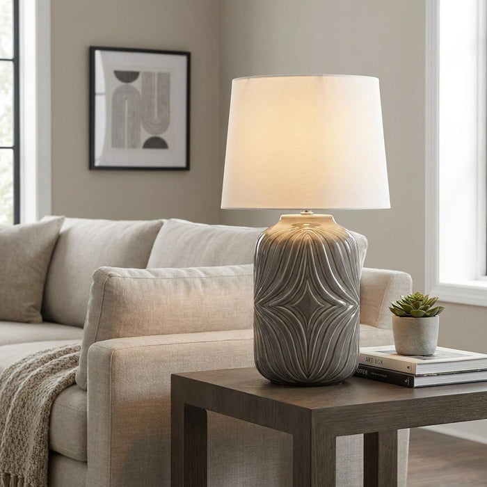 A lit Elstead Muse grey ceramic table lamp with a textured geometric base on a side table in a living room.