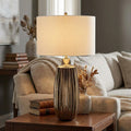 Bronze ceramic table lamp with a ribbed base and white drum shade sitting on a wooden side table in a living room.