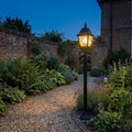 Black lantern lamp post illuminating a gravel garden path beside lavender plants and brick walls at dusk.