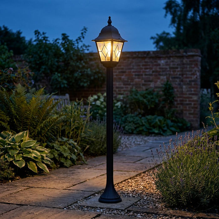 Black traditional lantern bollard light with leaded glass panels illuminating a garden stone path at dusk.