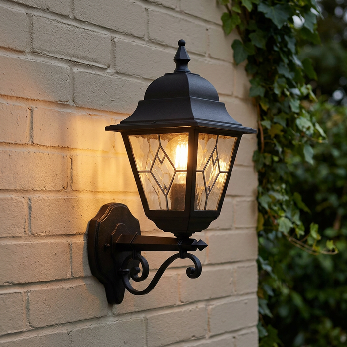Black traditional outdoor uplighter with leaded glass panels mounted on a brick wall, glowing warmly.