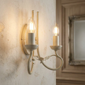 Ivory and gold 2-light wall sconce with elegant leaf motifs and glowing candle bulbs on a rustic, cracked plaster wall.