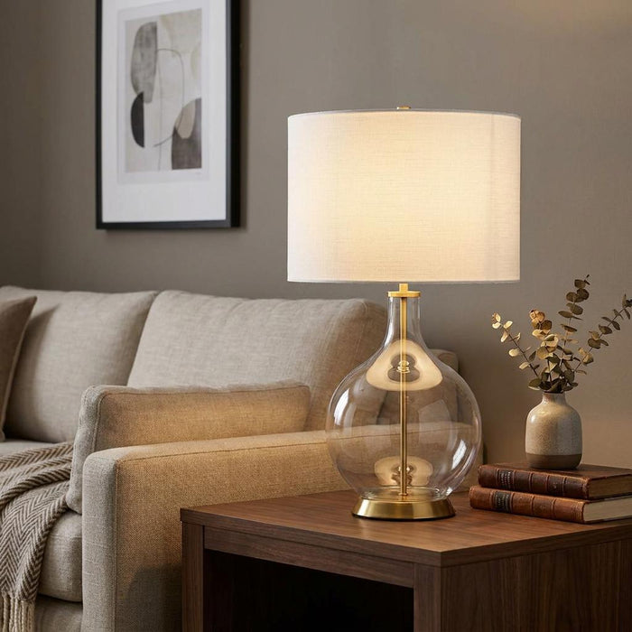 Brass and clear glass table lamp with a white drum shade lit on a wooden side table in a cozy living room.