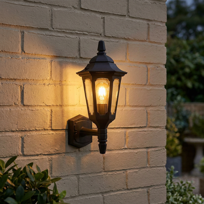A black traditional lantern wall light illuminates a pale brick wall with a warm, amber glow.