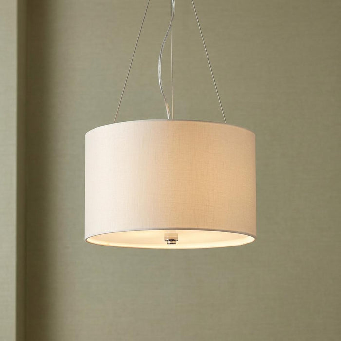 A lit cylindrical cream fabric drum pendant light suspended by thin wires against a neutral wall.
