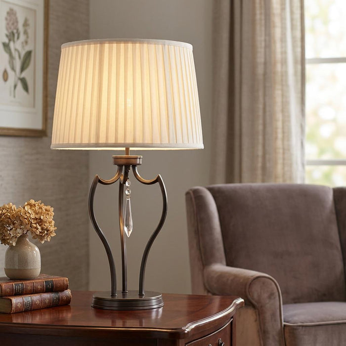 Dark bronze table lamp with a curved base, crystal droplet, and cream pleated shade on a wooden table.