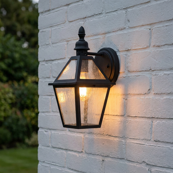 A black lantern-style wall light with seeded glass panels, lit from within and mounted on a white brick wall.