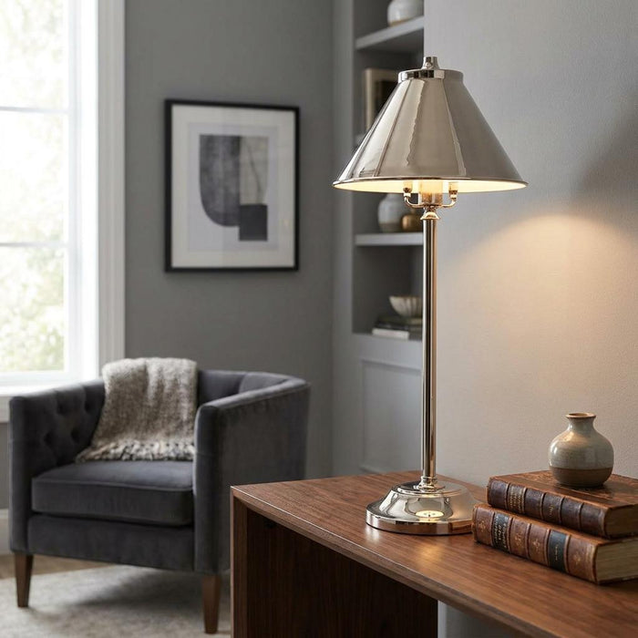 Polished nickel stick lamp with a conical shade lit on a wooden desk in a stylish grey living room.
