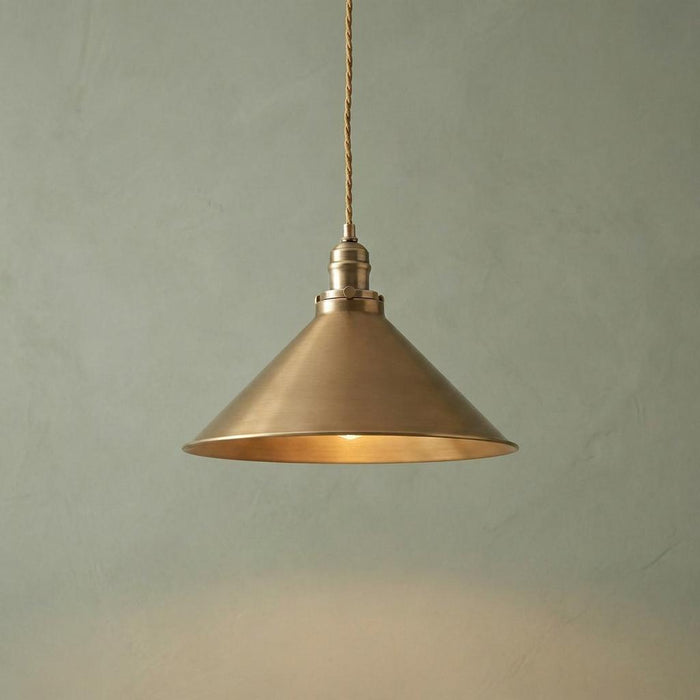 Aged brass conical pendant light hanging from a braided cord against a textured pale green wall.