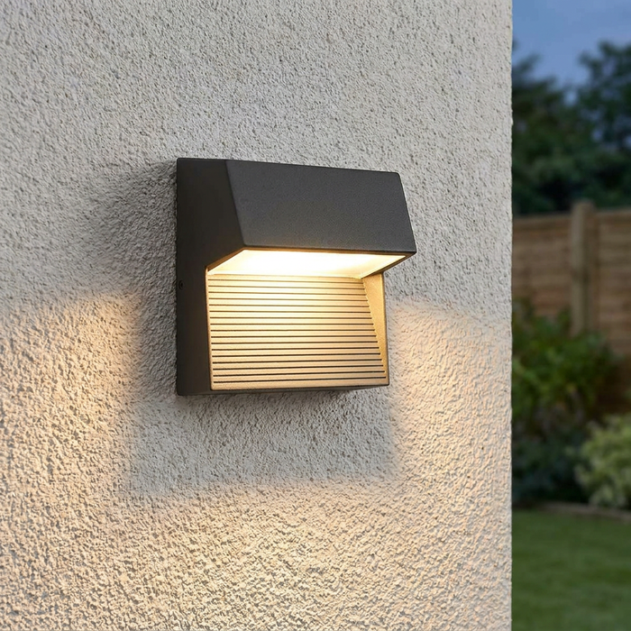 Graphite outdoor wall light with an illuminated slatted design mounted on a white textured wall.