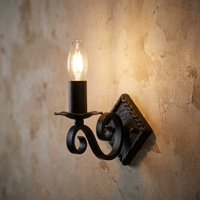 Black wrought iron candle-style wall sconce with a curved arm and lit bulb against a textured rustic wall.