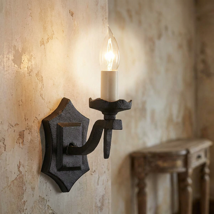 Wrought-iron wall sconce with a glowing candle bulb against a rustic, distressed plaster wall.