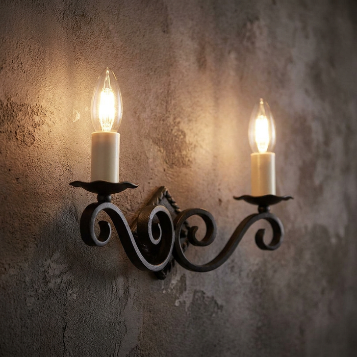 Black wrought iron wall sconce with scrollwork and two lit candle bulbs on a rustic textured wall.