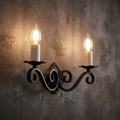 Black wrought iron wall sconce with scrollwork and two lit candle bulbs on a rustic textured wall.