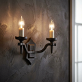 Two-light black wrought iron wall sconce with glowing candle-style bulbs on a dark textured wall.