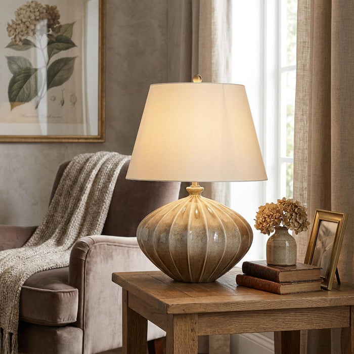 Ribbed pumpkin ceramic table lamp with a white shade glowing on a wooden side table in a cozy living room.