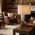 Bronze elephant table lamp with a linen shade on a side table in a cozy library with a fireplace.