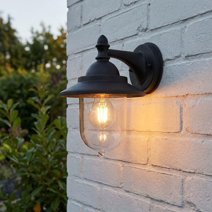 Black Elstead Sandown outdoor wall light with a warm glowing bulb, mounted on a white brick wall.