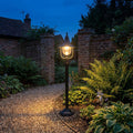 Black outdoor bollard light illuminating a gravel garden path and lush foliage next to brick walls at dusk.