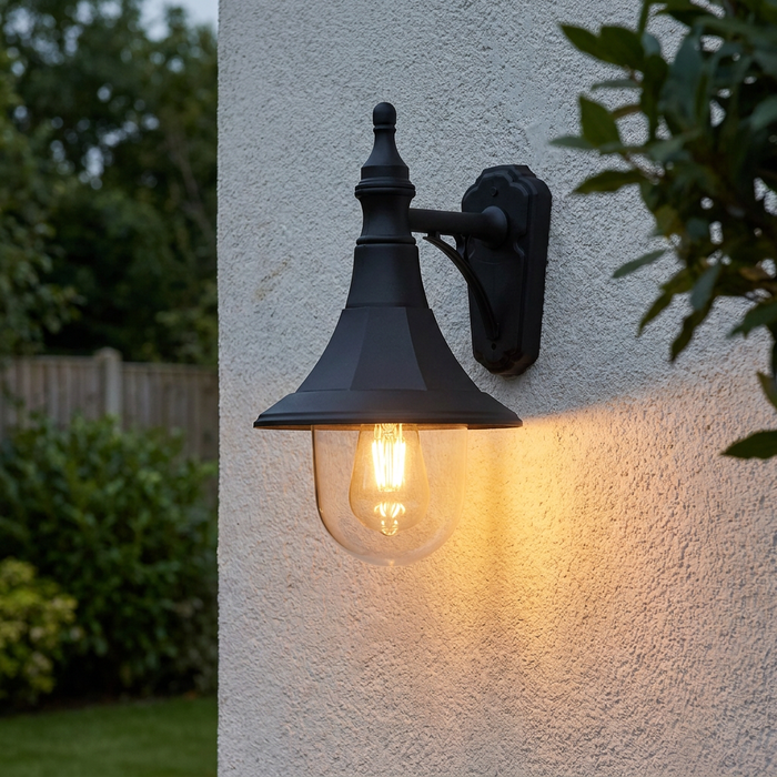 Traditional black outdoor downlighter wall light with a warm glowing filament bulb mounted on a textured white wall.