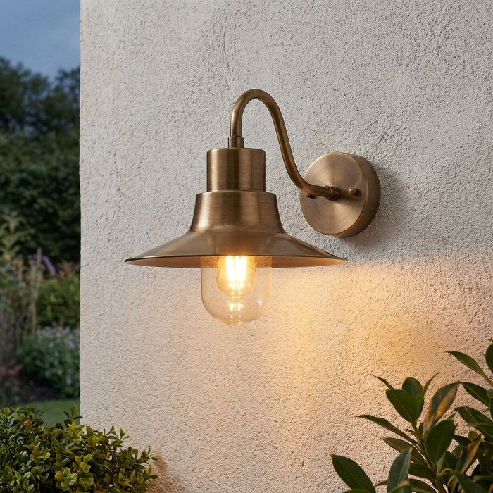 Burnished brass outdoor wall light with a wide conical shade and glass cover, mounted on a textured white wall.