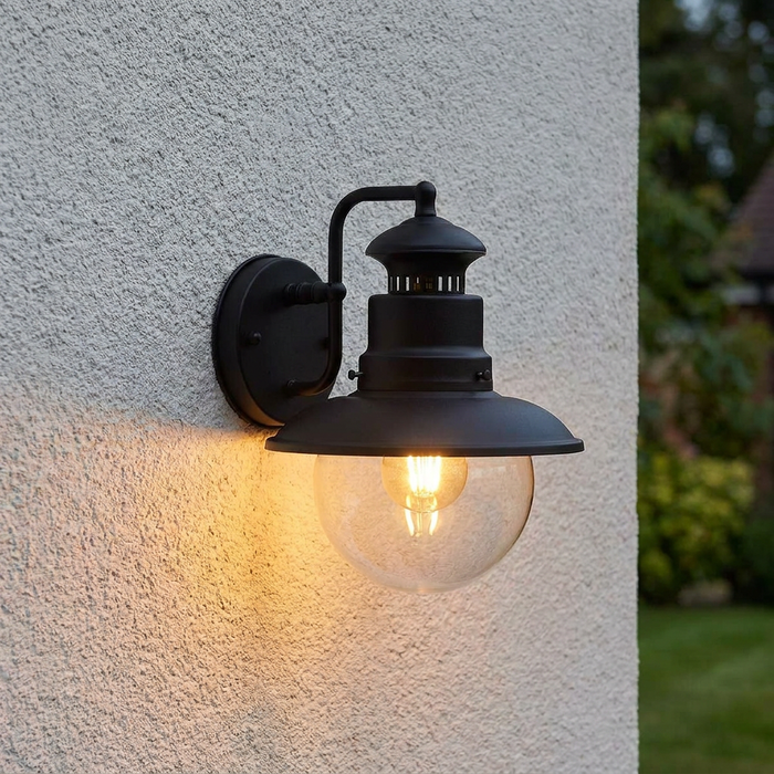 Black outdoor wall lantern with a clear glass globe and warm glowing bulb mounted on a textured white wall.