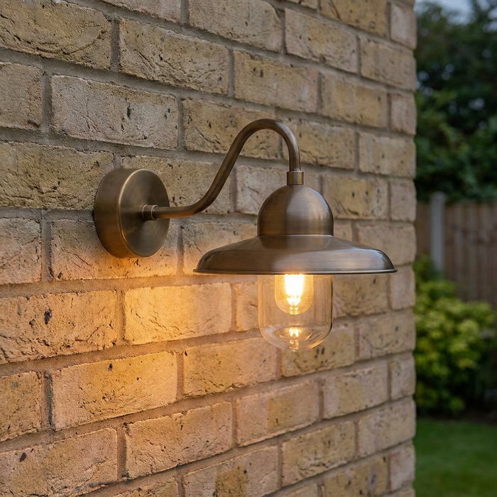 Antique brass outdoor wall light with swan neck arm and clear glass shade illuminated on a yellow brick wall.
