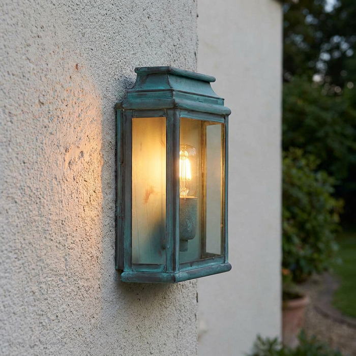 Verdigris outdoor wall lantern with a glowing filament bulb mounted on a textured white wall.