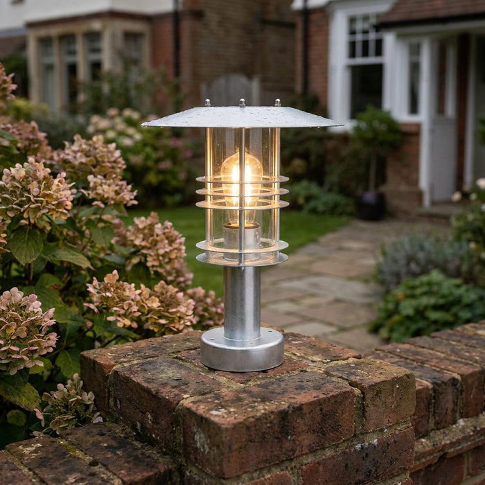 Galvanized steel pedestal lantern with a warm Edison bulb, mounted on a brick garden wall.