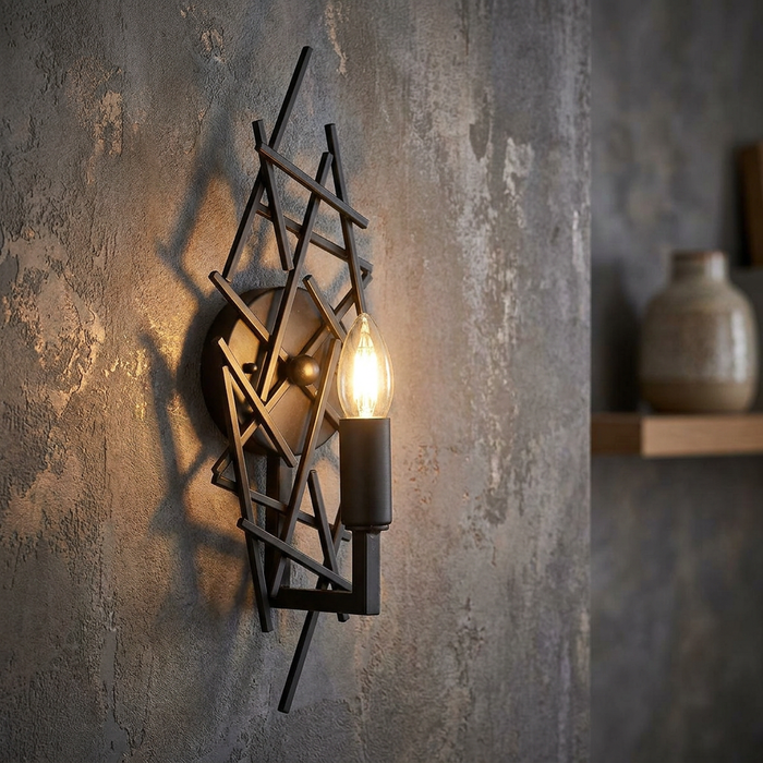 Modern bronze wall light featuring geometric crisscrossing rods and a lit candle bulb on a textured grey wall.