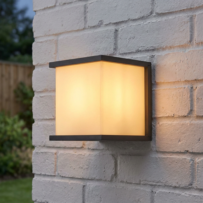 Modern square graphite outdoor wall light with a warm glow mounted on a white brick wall.