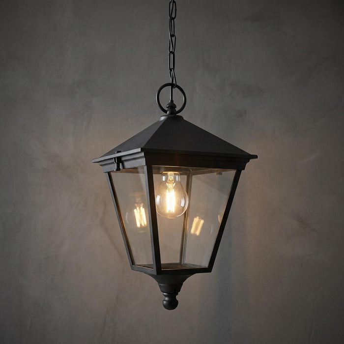 Traditional black outdoor pendant lantern with clear glass panels and a lit bulb hanging against a textured grey wall.