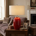 Red ceramic table lamp with a white drum shade on a wooden side table in a living room setting.