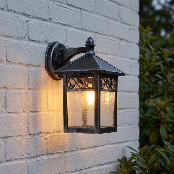 Elstead Winchcombe outdoor wall lantern with black/silver finish and leaf detail, glowing on a white brick wall.