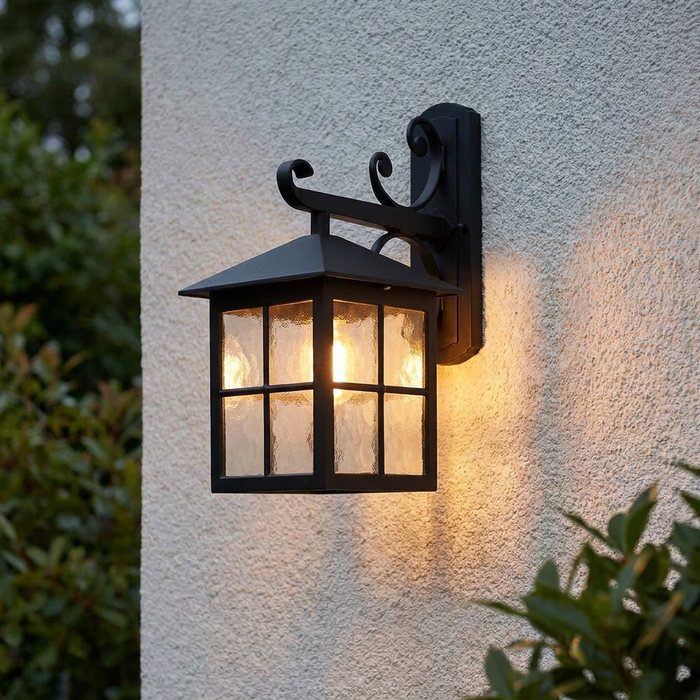 A traditional black outdoor lantern with textured glass panes, emitting a warm glow on a white textured wall.