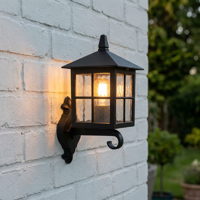 A black Elstead Winchester outdoor lantern wall light with a glowing bulb, mounted on a white brick wall.
