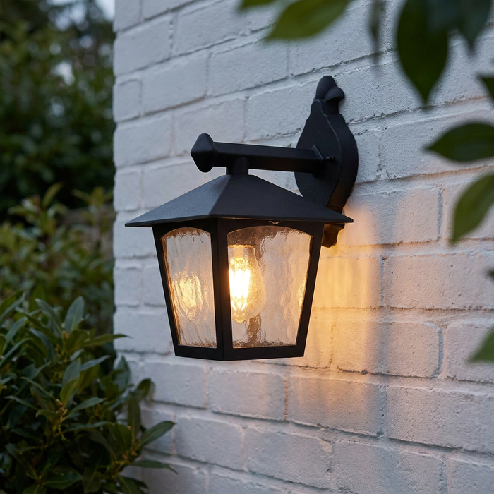 A black outdoor lantern with textured glass mounted on a white brick wall, casting a warm glow from within.