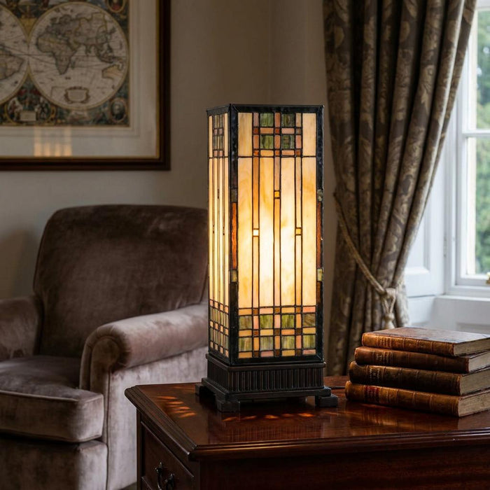 Tall illuminated Empire Tiffany square table lamp with geometric stained glass on a desk with vintage books.